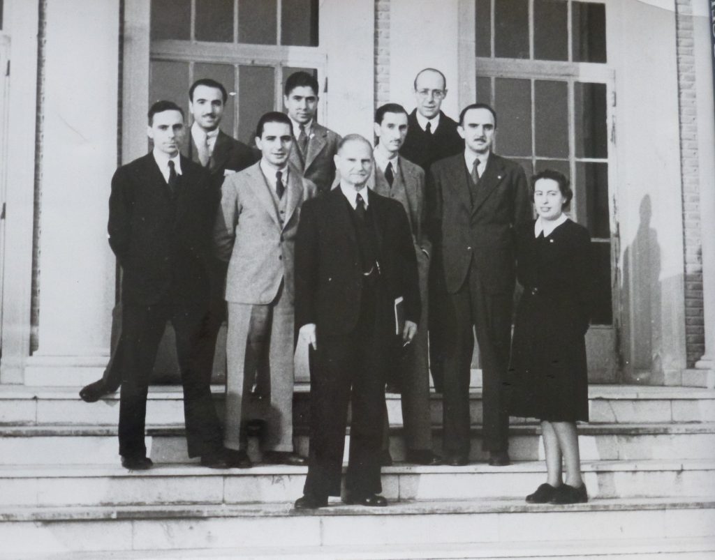 FFMTV-FDE-001 Old black and white photo in which 7 men in suits are seen on a staircase behind another older man holding a book. To one side, a woman on the same step as the older man