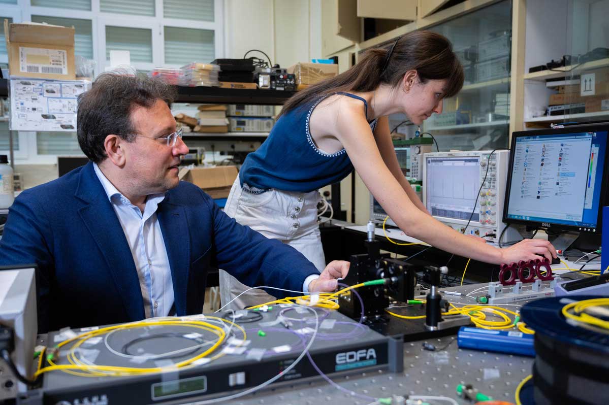 juan_diego_ania-25-Ines-Caceres-med En un laboratorio un hombre con americana azul está sentado a la izquierda mientras mira a una mujer joven a la derecha que opera de pie un ordenador