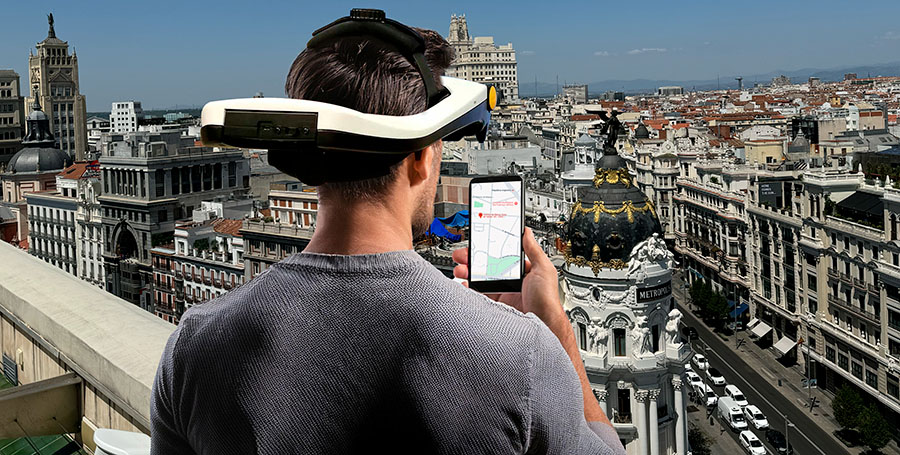 Nuevo montaje gafas gran via-900px Un hombre joven de espaldas lleva puesto un casco de simulación Simvis, mientras mira de cerca un móvil y ve también de lejos la calle Gran Vía de Madrid