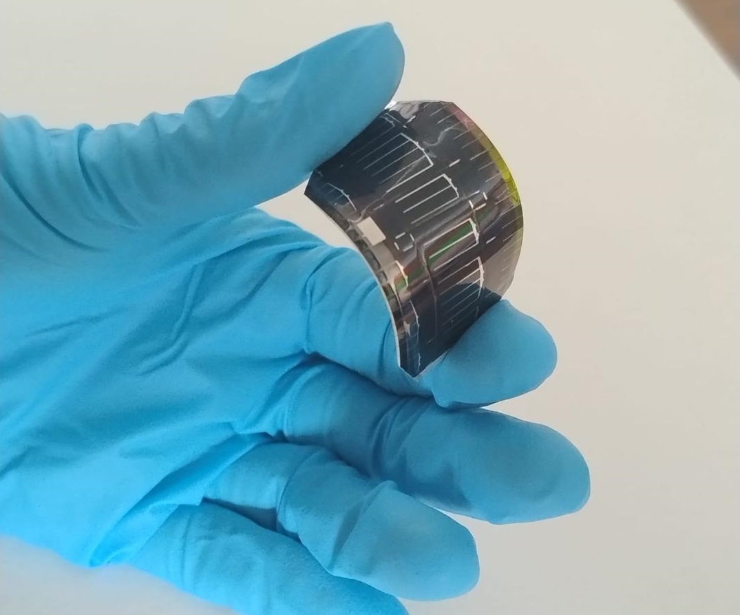 Muestra de célula-SUNLIFE Una mano con un guante de laboratorio sujeta una célula solar en forma de lámina flexible oscura